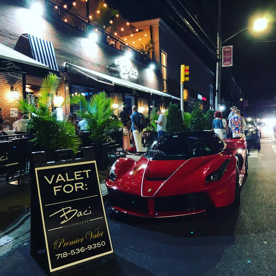 Red Ferrari at the curb beside a Baci Ristorante valet sign at night, with diners seated outside