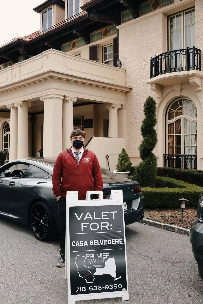 Valet attendant in a red jacket and mask behind a Premier Valet sign for Casa Belvedere