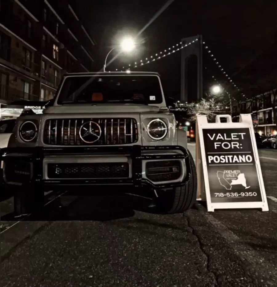 Mercedes G-Class parked at night beside a Valet For: Positano sign, with a lit suspension bridge behind