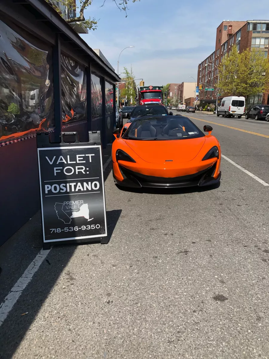 Orange McLaren convertible at the curb beside a Valet For: Positano sign on a sunny city avenue