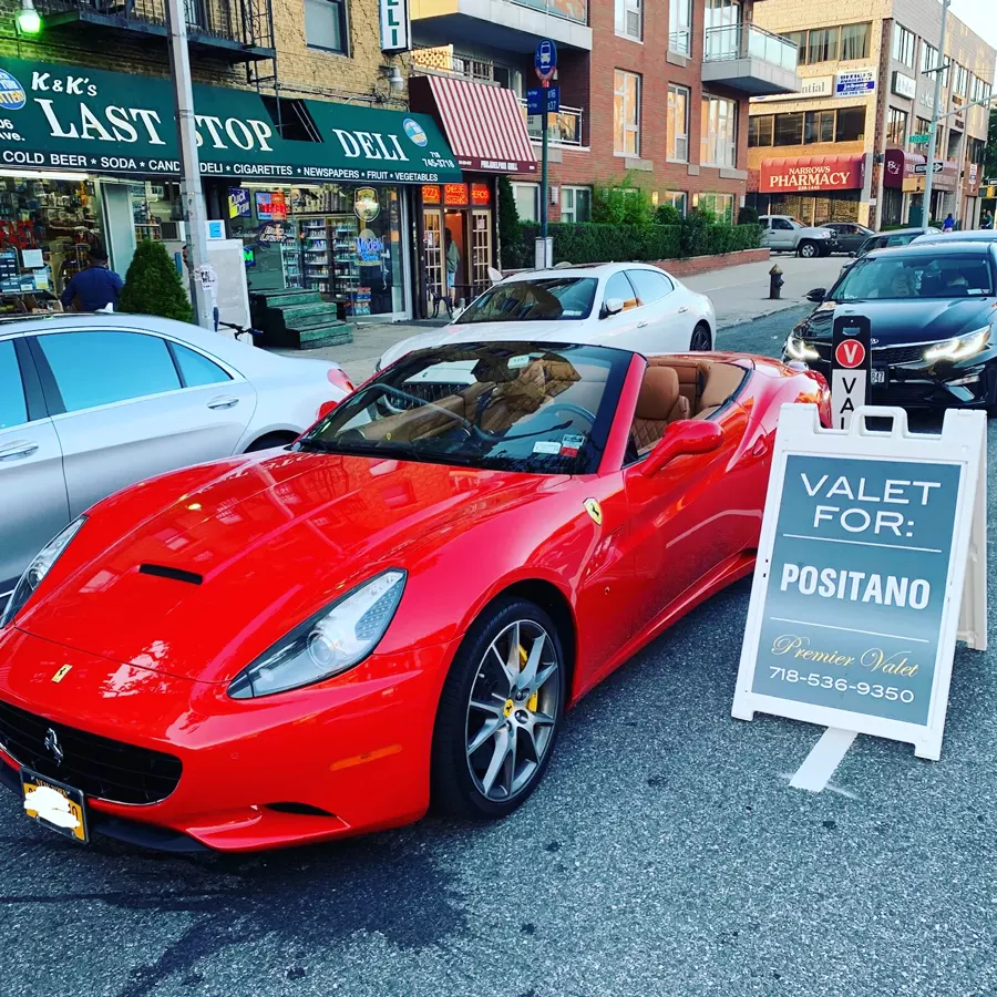 Red Ferrari convertible with tan interior parked beside a Valet For: Positano sign in front of a deli
