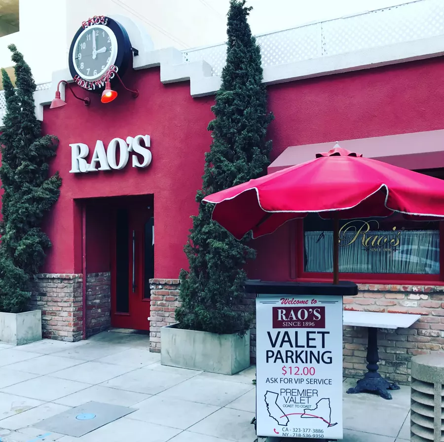 Valet stand under a red umbrella outside Rao's red facade, below a clock reading Rao's Hollywood