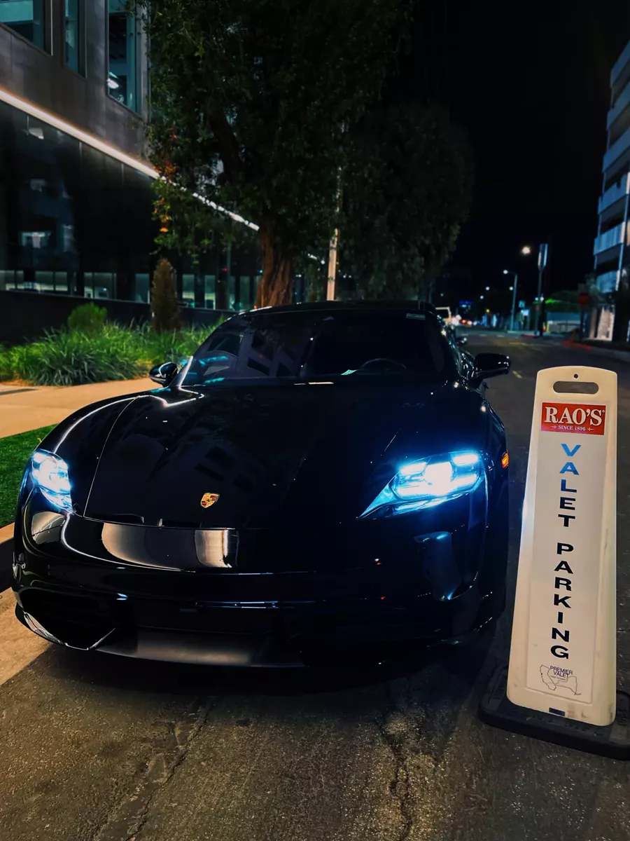 Black Porsche with headlights on parked beside a Rao's valet parking cone at night