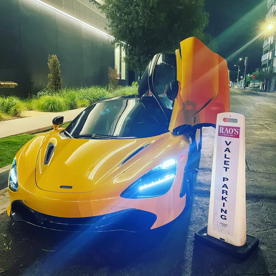 Orange McLaren with its door raised beside a Rao's valet parking cone at night