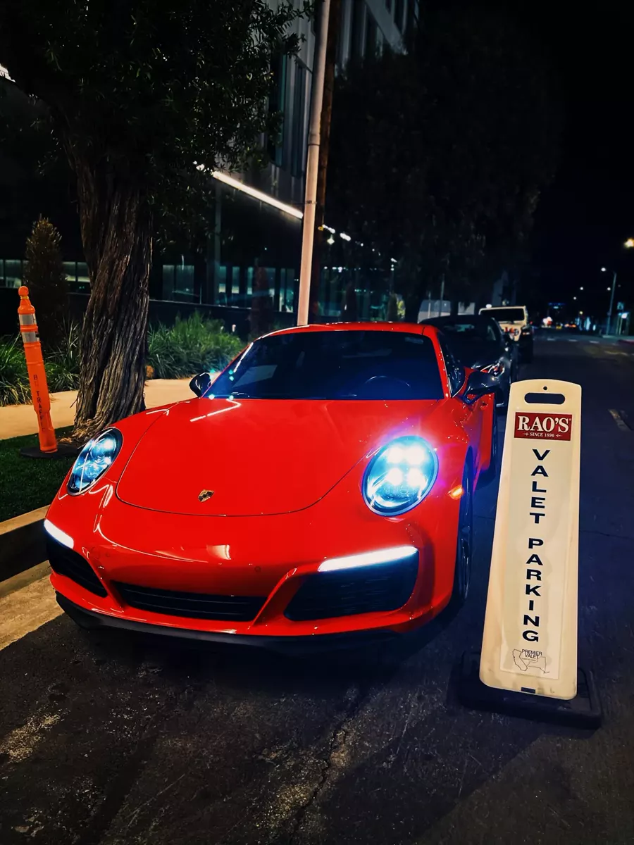 Red Porsche with headlights on parked beside a Rao's valet parking cone at night