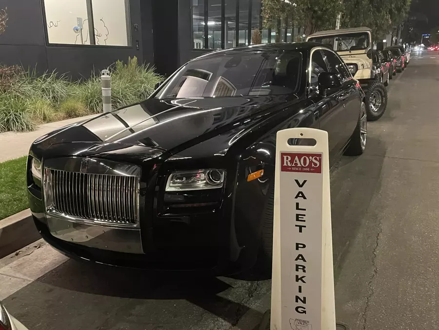 Black Rolls-Royce parked beside a Rao's valet parking cone, with a line of cars behind it at night