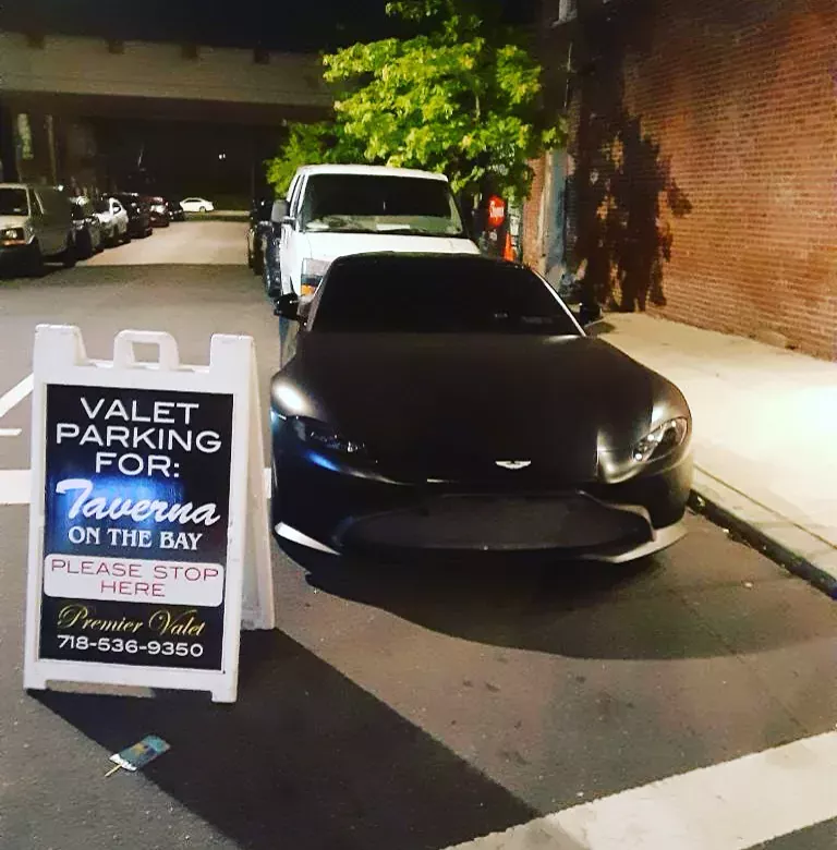 Premier Valet sign for Taverna On The Bay beside a matte black Aston Martin at night