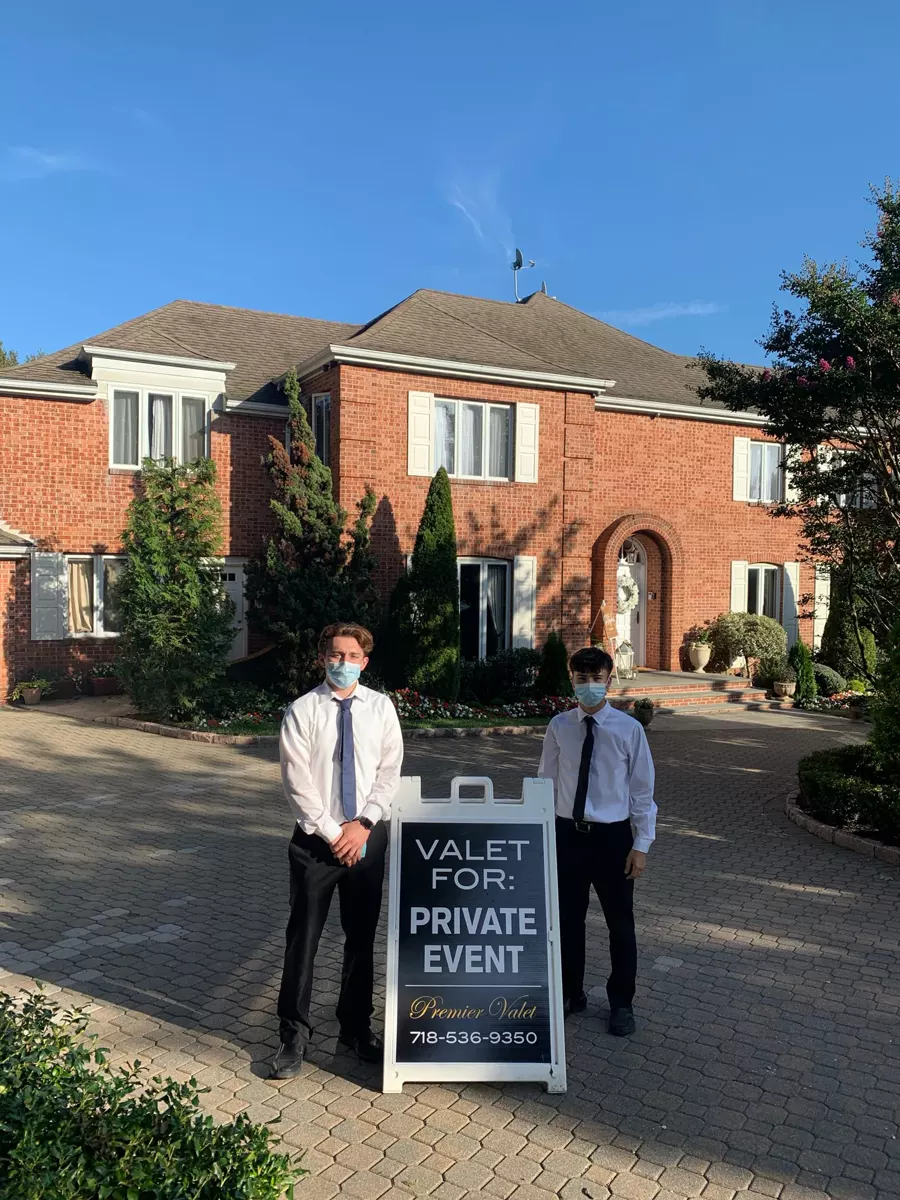 Two masked valet attendants beside a Premier Valet private event sign in front of a brick house