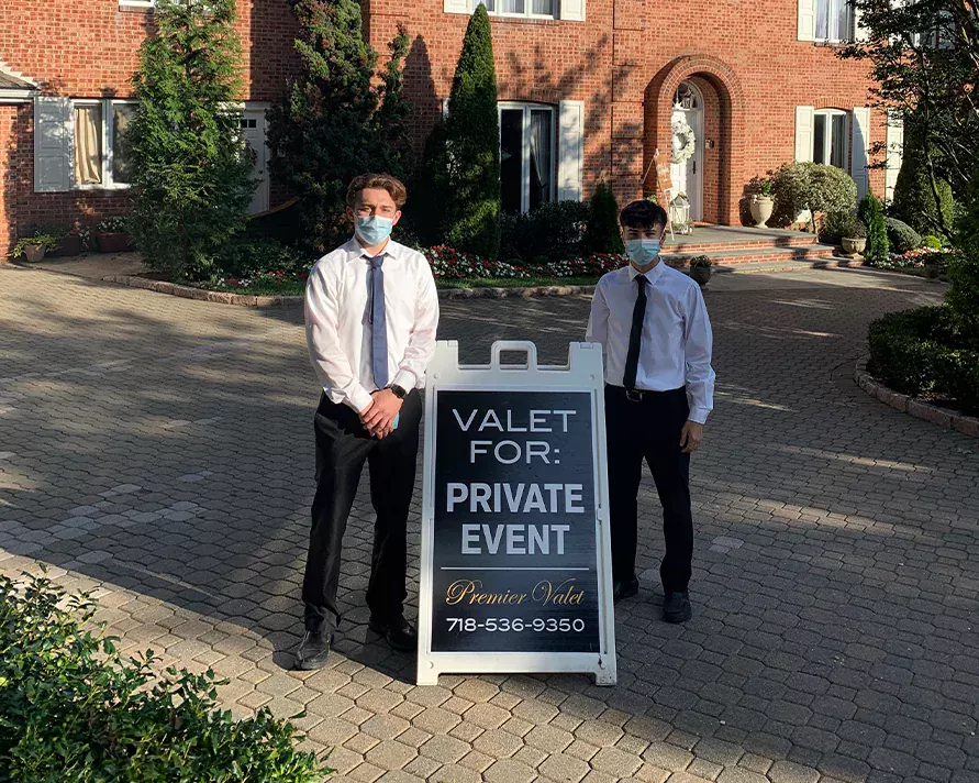 Two masked valet attendants flanking a Premier Valet private event sign on a brick-paved driveway