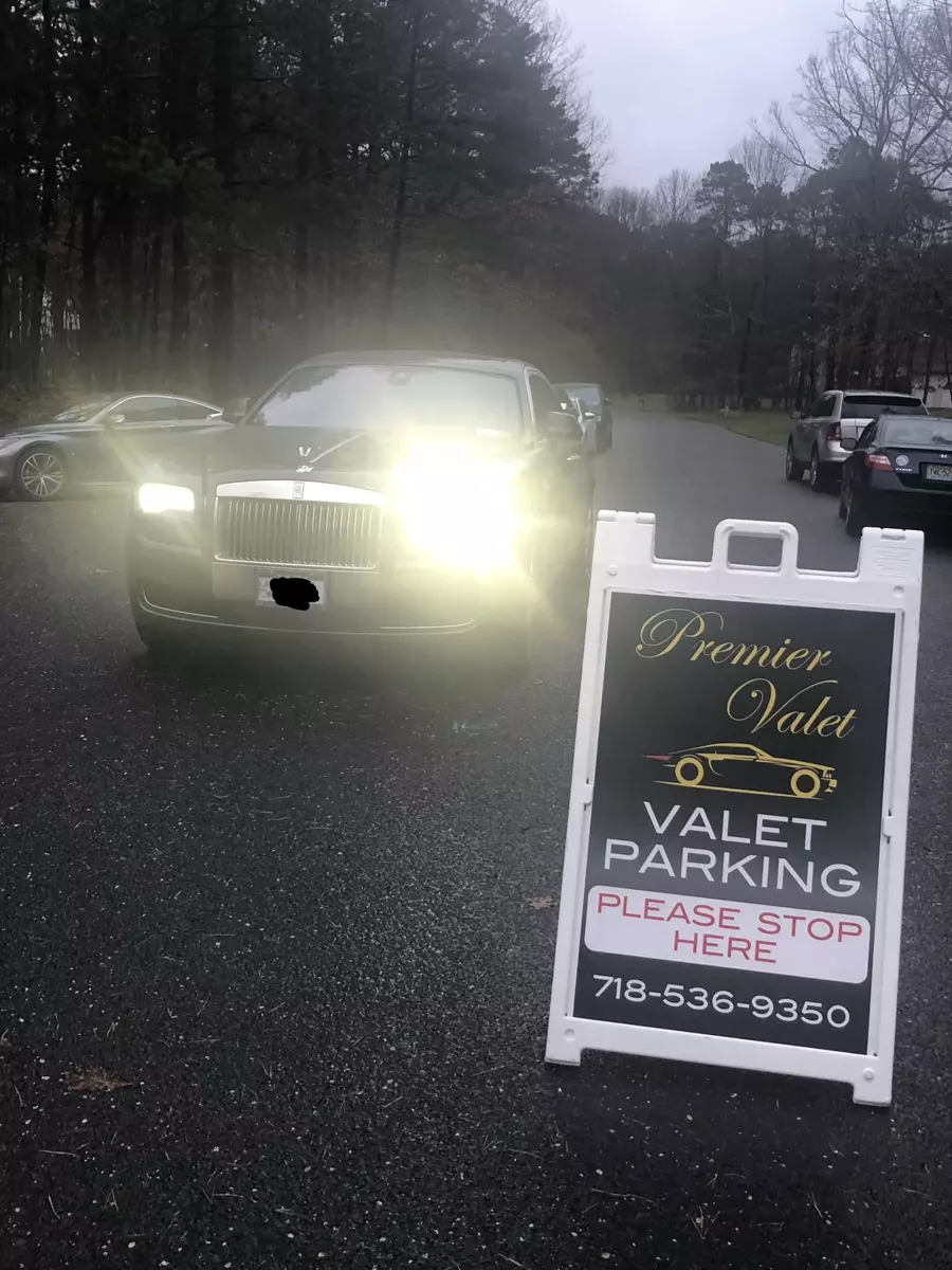 Black Rolls-Royce with headlights on beside a Premier Valet parking sign on a tree-lined road at dusk