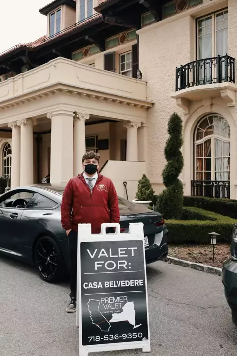 Valet attendant in a red jacket and mask behind a Premier Valet sign for Casa Belvedere