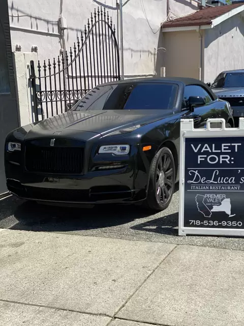 Black Rolls-Royce convertible by an iron gate beside a Premier Valet sign for DeLuca's Italian Restaurant