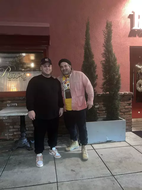 Two men posing on a sidewalk in front of a red wall and a window lettered Rao's Since 1896