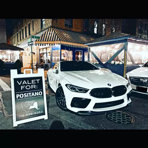 White BMW coupe parked at night beside a Valet For: Positano sign outside a restaurant with outdoor dining