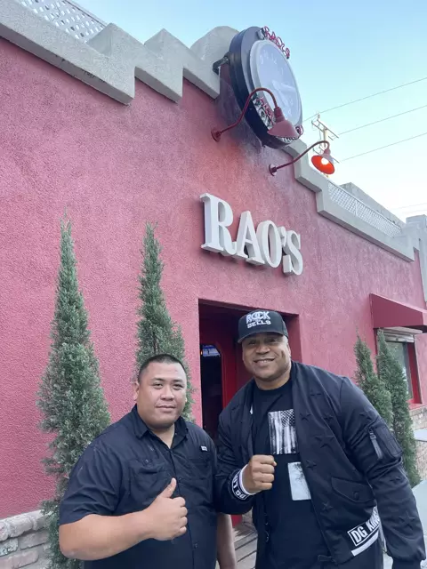 Two men posing outside a red building with a white RAO'S sign and a large wall clock