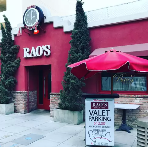 Valet stand under a red umbrella outside Rao's red facade, below a clock reading Rao's Hollywood