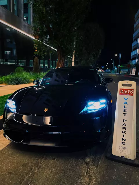 Black Porsche with headlights on parked beside a Rao's valet parking cone at night