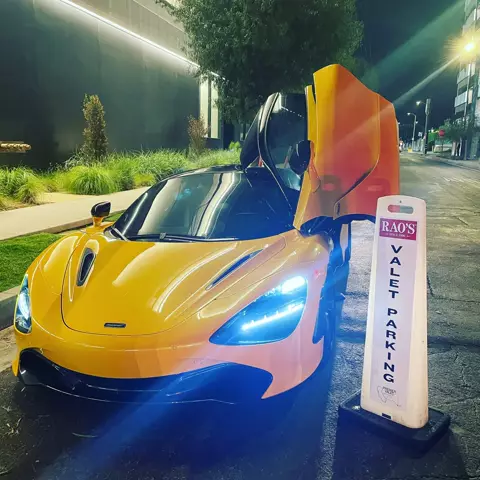 Orange McLaren with its door raised beside a Rao's valet parking cone at night
