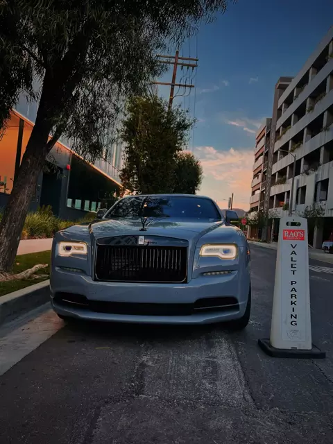 Grey Rolls-Royce facing the camera beside a Rao's valet parking cone under a sunset sky