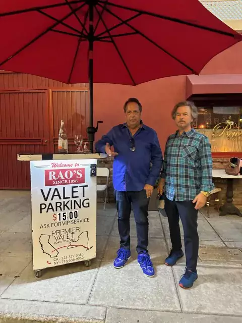 Two men standing beside a Rao's valet parking podium under a red umbrella