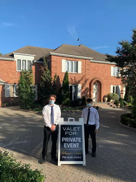 Two masked valet attendants beside a Premier Valet private event sign in front of a brick house