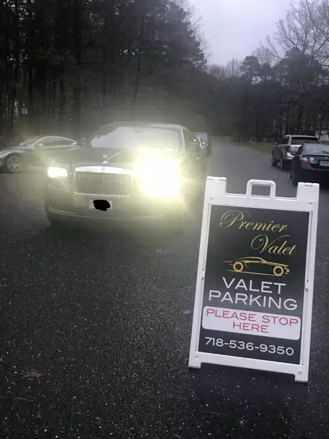 Black Rolls-Royce with headlights on beside a Premier Valet parking sign on a tree-lined road at dusk