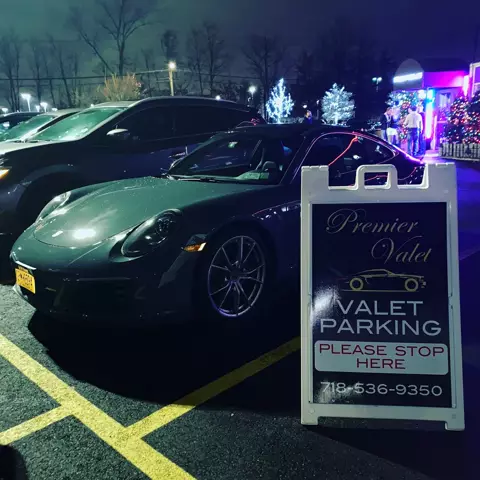Dark gray Porsche 911 beside a Premier Valet sign reading Please Stop Here, with holiday lights behind